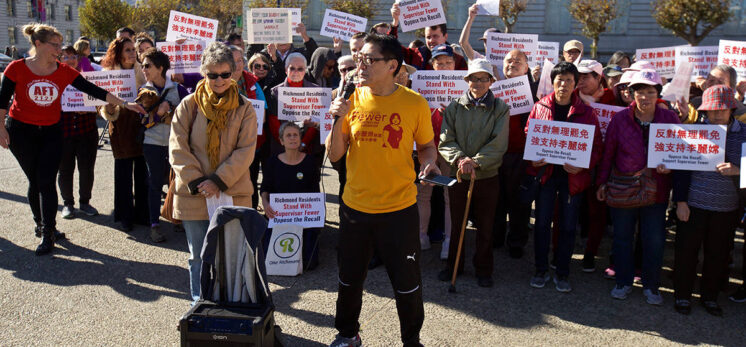 Crowd of people holding up signs in an Asian language protest with Don Misumi of Richmond Rising in the front with a yellow shirt and microphone