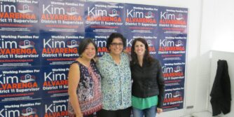 Three women (L-R: Sandra Lee Fewer, Kimberly Alvarenga, and Hillary Ronen) stand in front of a "selfie wall" covered with Kim Alvarenga campaign signs