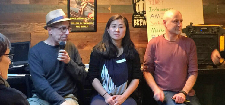 A man with a microphone, a woman, and another man seated at a community event at a bar