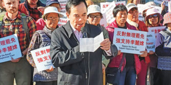 Protestors holding signs in an Asian language, with a man in the front reading from a sheet of paper